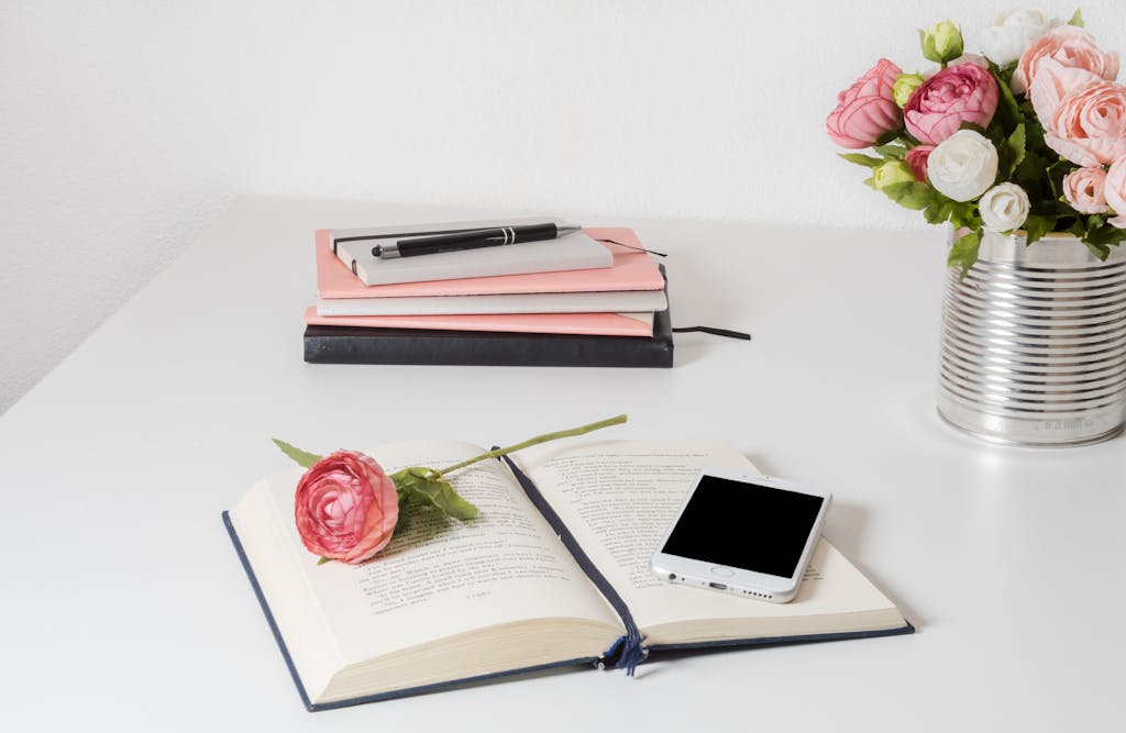 A stylish home office setup with an open book, smartphone, and flowers on a white desk.